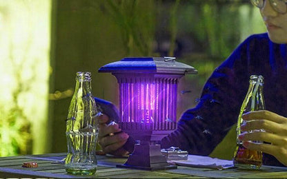 Lampe anti moustique JARDIN KILLERS émettant une lumière violette, idéale pour l'extérieur, avec des bouteilles à proximité.